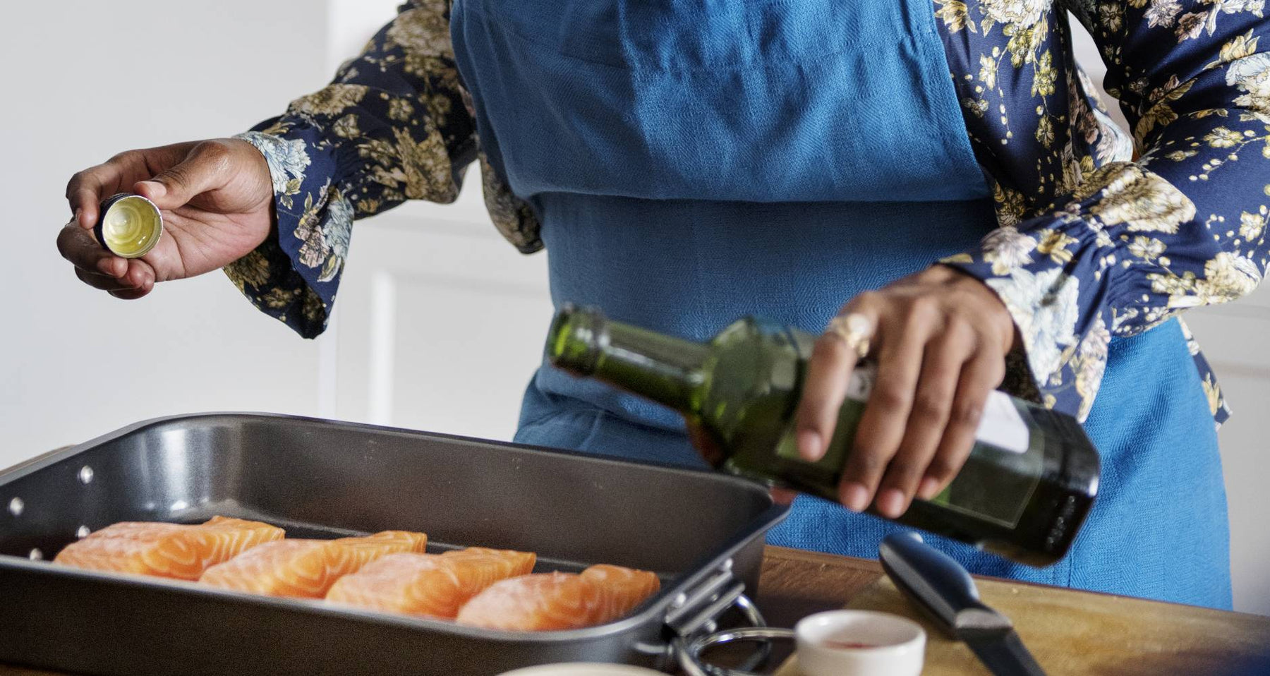 woman in a blue apron seasoning bright orange, raw salmon in a black baking dish
