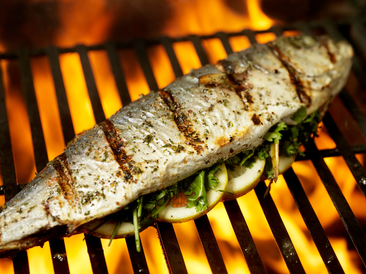 a small european sea bass with defined grill marks, skin dappled with salt and pepper, stuffed with herbs and citrus on top of a grill with high orange and red flames. 